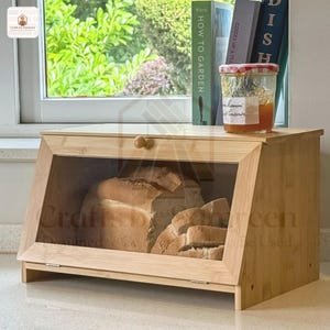 Puede incluir: Una panera de bambú con frente de cristal transparente, que muestra pan rebanado. La panera tiene un pomo de madera y una parte superior inclinada. El texto "Crafts by Sunmoon" es visible.