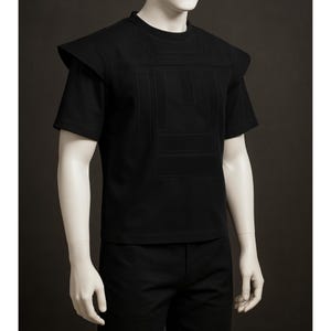 以下が含まれることがあります： 前面に幾何学模様と肩パッドが付いた黒いTシャツ。半袖のシャツは無地の黒い素材で作られています。シャツはマネキンに展示されています。