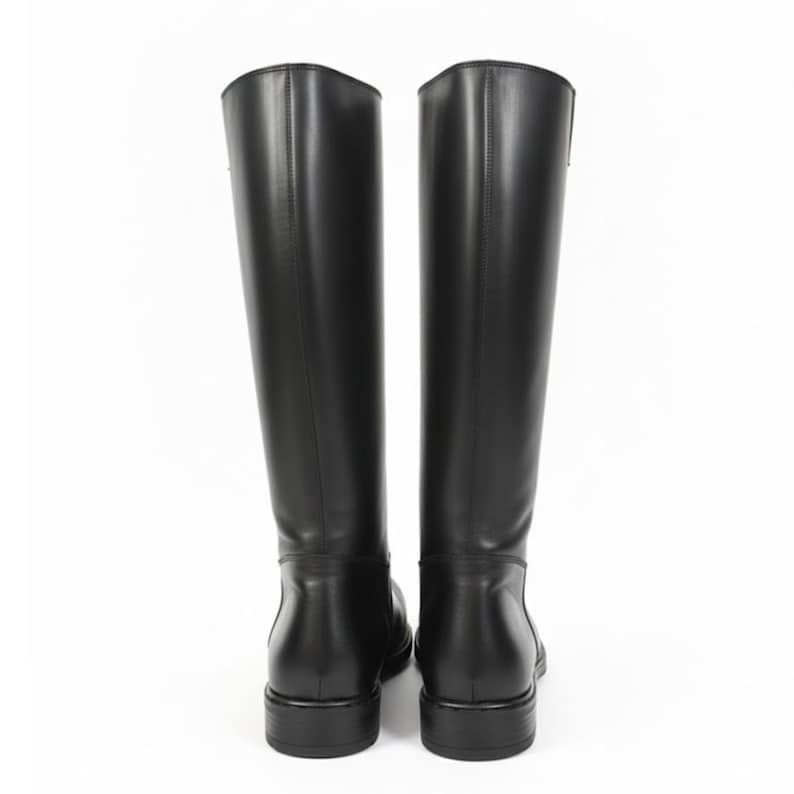 May include: A pair of knee-high black boots with a smooth finish, featuring a structured shaft, a rounded toe, and a low block heel. The boots are shown from the back view, displaying vertical seam detailing along the center of the shaft.