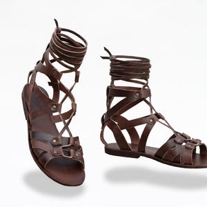 Puede incluir: Sandalias gladiadoras de cuero marrón con múltiples correas y diseño de cordones. Las sandalias tienen suela plana y están diseñadas para envolver el tobillo y la parte inferior de la pierna. Fondo blanco.