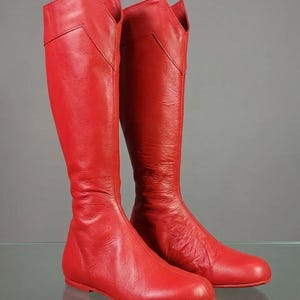 K&ouml;nnte beinhalten: Ein Paar hohe, rote Lederstiefel. Die Stiefel haben eine flache Sohle und ein spitzes, eckiges Design am oberen Schaft. Die Stiefel sind kniehoch und aus glattem, gl&auml;nzendem Leder gefertigt.