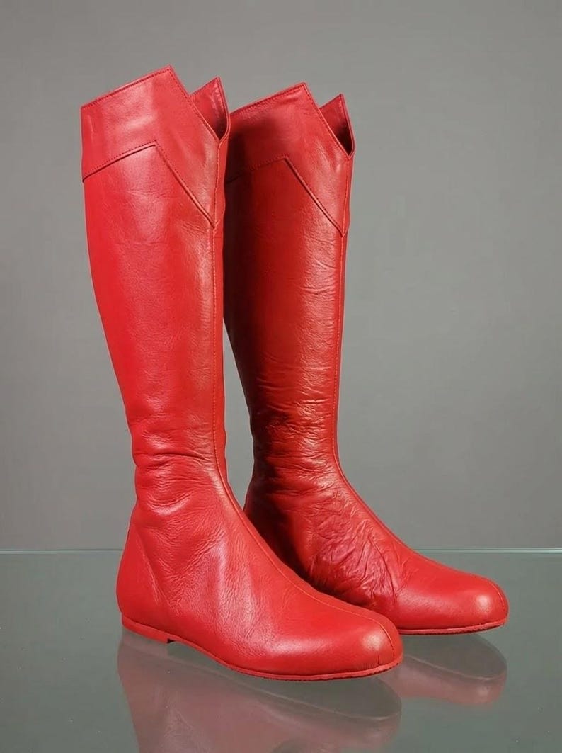 K&ouml;nnte beinhalten: Ein Paar hohe, rote Lederstiefel. Die Stiefel haben eine flache Sohle und ein spitzes, eckiges Design am oberen Schaft. Die Stiefel sind kniehoch und aus glattem, gl&auml;nzendem Leder gefertigt.