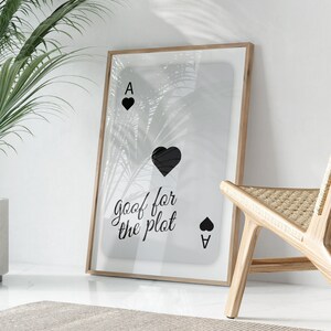 Puede incluir: Una impresión enmarcada de una carta de juego con un as de picas negro y el texto "goof for the plot" en fuente cursiva negra. La carta se superpone a una imagen borrosa de hojas de palma.