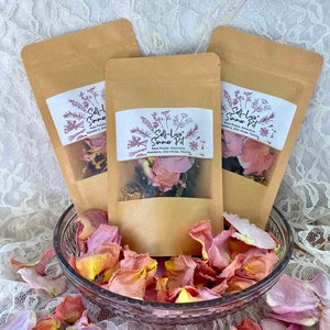 Può includere: Tre sacchetti di carta marrone con etichette bianche che recitano "Self-Love Summer Pot" e elencano gli ingredienti: petali di rosa, calendula, rosmarino, anice stellato, vaniglia. I sacchetti sono pieni di fiori e erbe secche. I sacchetti sono su un tovagliolo di pizzo bianco con una ciotola di vetro piena di petali di rosa.