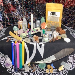Puede incluir: Un paño negro con un diseño de pentagrama plateado, rodeado de varios cristales, velas, conchas, plumas y una pequeña bolsa etiquetada "Dream Tea".