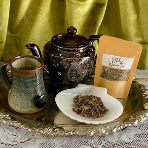 May include: A silver tray with a brown teapot, a mug of tea, and a small bag of loose leaf tea. The tea bag is labeled "Witchy Woman Tea".
