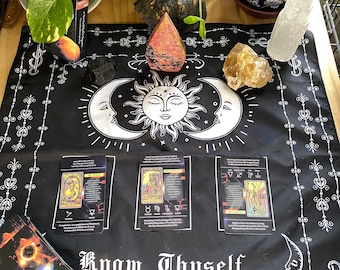 May include: Black and white tarot cloth with a sun and moon design. The cloth has the words "Know Thyself" printed on it. There are three tarot cards laid out on the cloth, along with a variety of crystals and other objects.