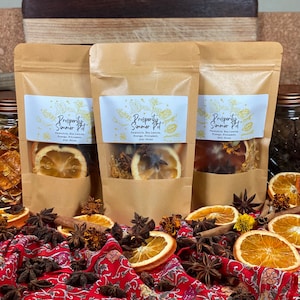 May include: Three brown paper bags with clear windows, each containing a mix of dried orange slices, star anise, cinnamon sticks, and bay leaves. The bags are labeled "Prosperity Summer Pot" and list the ingredients. The bags are sitting on a red and white patterned fabric with more dried orange slices, star anise, and cinnamon sticks scattered around.