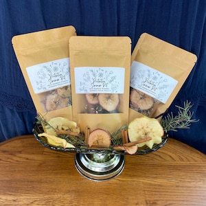 May include: Three brown paper bags with white labels that say "Protection Summer Pot" with a list of ingredients. The bags are sitting on a silver cake stand with dried apple slices, cinnamon sticks, and rosemary sprigs.