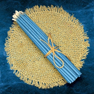 May include: A bundle of blue taper candles tied with twine, resting on a round, fringed, golden-brown jute placemat. The background is a dark blue, textured surface.