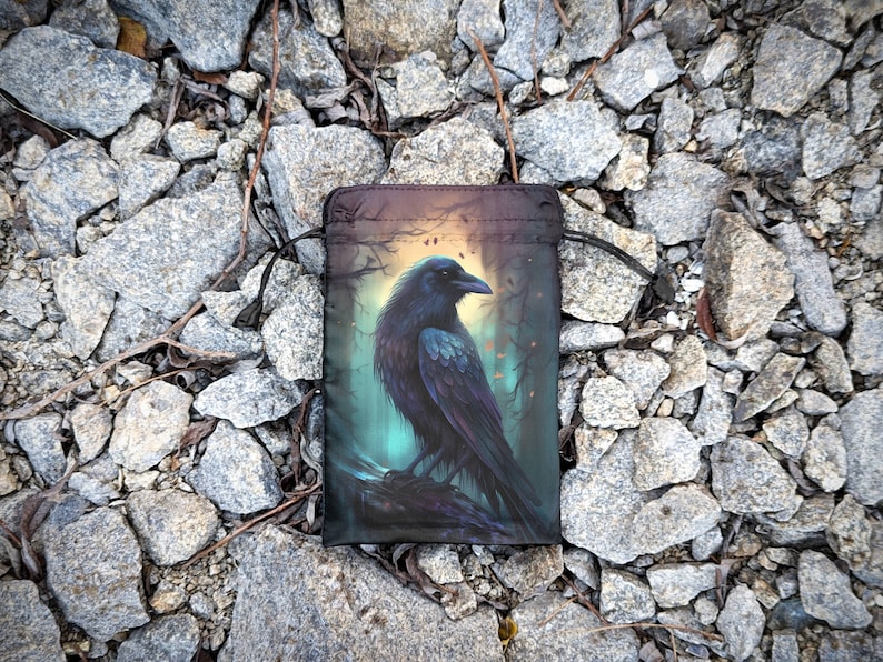 May include: A black drawstring bag with a raven illustration. The raven is perched on a branch in a dark forest setting with a glowing light behind it.