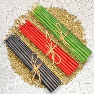 May include: Three bundles of candles, one black, one red, and one green, are tied together with twine. The candles are arranged on a gold-colored surface.