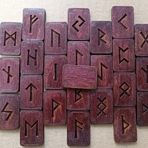 May include: A set of 25 wooden runes with a purple-brown stain. Each rune is engraved with a different symbol. The runes are arranged in a rectangular pattern.