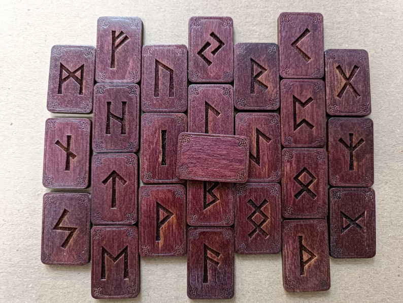 May include: A set of 25 wooden runes with a purple-brown stain. Each rune is engraved with a different symbol. The runes are arranged in a rectangular pattern.