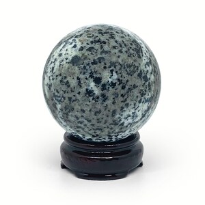 Può includere: Una sfera grigia maculata, forse una pietra decorativa o una palla di cristallo, poggia su un supporto rotondo in legno marrone scuro. La sfera ha un aspetto screziato con macchie grigie scure e chiare. Il supporto ha una finitura lucida.