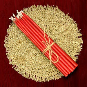 May include: A bundle of red taper candles tied together with twine, resting on a round, crocheted placemat with a fringed edge.