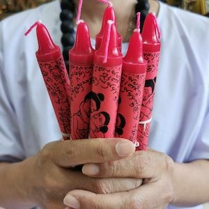 Può includere: Un mazzo di candele rosa con illustrazioni in bianco e nero di persone e testo in una lingua straniera. Le candele sono tenute nelle mani di una persona.