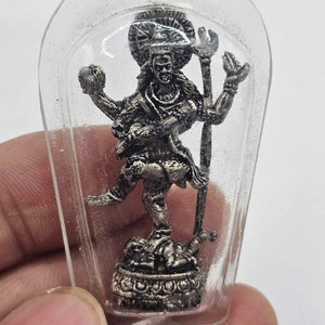 May include: A silver-colored figurine of a deity, enclosed in a clear plastic case. The figure has multiple arms and is posed in a dynamic stance, holding various objects. The base of the figurine features intricate details.