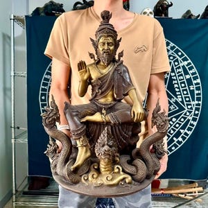 May include: A bronze-colored statue of a seated figure with a beard, ornate headdress, and raised hand. The statue is flanked by two serpent-like figures and rests on a round base. The figure is wearing a robe and jewelry.