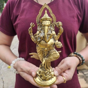 May include: A gold-colored brass statue of Ganesha, the Hindu god of wisdom and prosperity. The statue depicts Ganesha standing on a base with his trunk raised and his hands in a mudra. The statue is intricately detailed and has a traditional design.