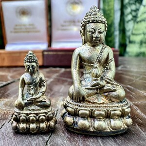 Può includere: Due antiche statue di Buddha color oro. La statua più grande è seduta su una base di loto dettagliata, con dettagli intricati sulla veste e sulla testa. La statua più piccola rispecchia quella più grande. Entrambe sono in posa meditativa.