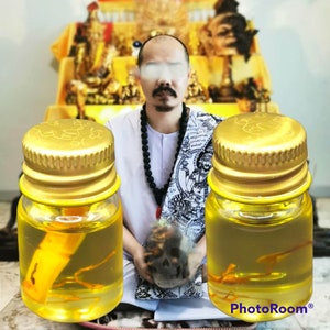 May include: Two small glass bottles with gold lids filled with a yellow liquid and containing small, dried, orange-colored objects. The bottles are in front of a man wearing a white shirt and a black beaded necklace.