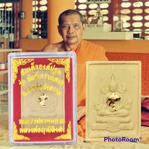 Peut inclure: Une amulette blanche avec des accents dorés représentant une figure de Bouddha assis. L'amulette est dans un boîtier en plastique transparent avec un fond rouge et des garnitures dorées. Le boîtier porte du texte thaïlandais sur le devant.