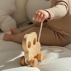 May include: A wooden pull toy shaped like an owl with a rope attached. The toy has a natural wood finish with engraved details. The owl toy is on a white surface. The toy is being held by a person wearing a tan ribbed onesie.