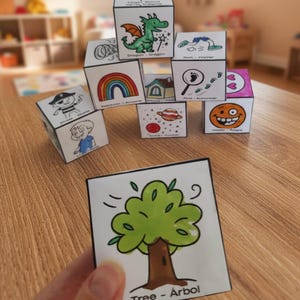 Puede incluir: Bloques ilustrados con varios dibujos, incluyendo un dragón, un arcoíris, una casa y una cara sonriente. Una mano sostiene una tarjeta con una ilustración de un árbol y el texto "Tree - Árbol". Los bloques están apilados.