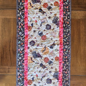 May include: A rectangular quilted table runner featuring a central panel with Halloween motifs including ghosts, black cats, bats, and pumpkins on a cream background, bordered by a bright pink strip and a dark outer border with candy patterns.