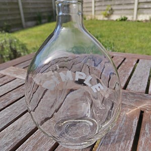 May include: Clear glass bottle with a rounded body and a narrow neck. The word "SIMPLE" is etched on the front. The bottle is on a wooden table outdoors.