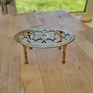 May include: An oval brass trivet featuring an intricate openwork floral pattern in the center. The piece is supported by four slender, cylindrical legs with subtle decorative rings near the top, providing a stable base for this metallic kitchen accessory.