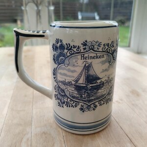 May include: A white ceramic beer stein with a blue and white floral design and the word "Heineken". The stein features a handle and a detailed illustration of a sailboat on the front. The stein is approximately 6 inches tall.