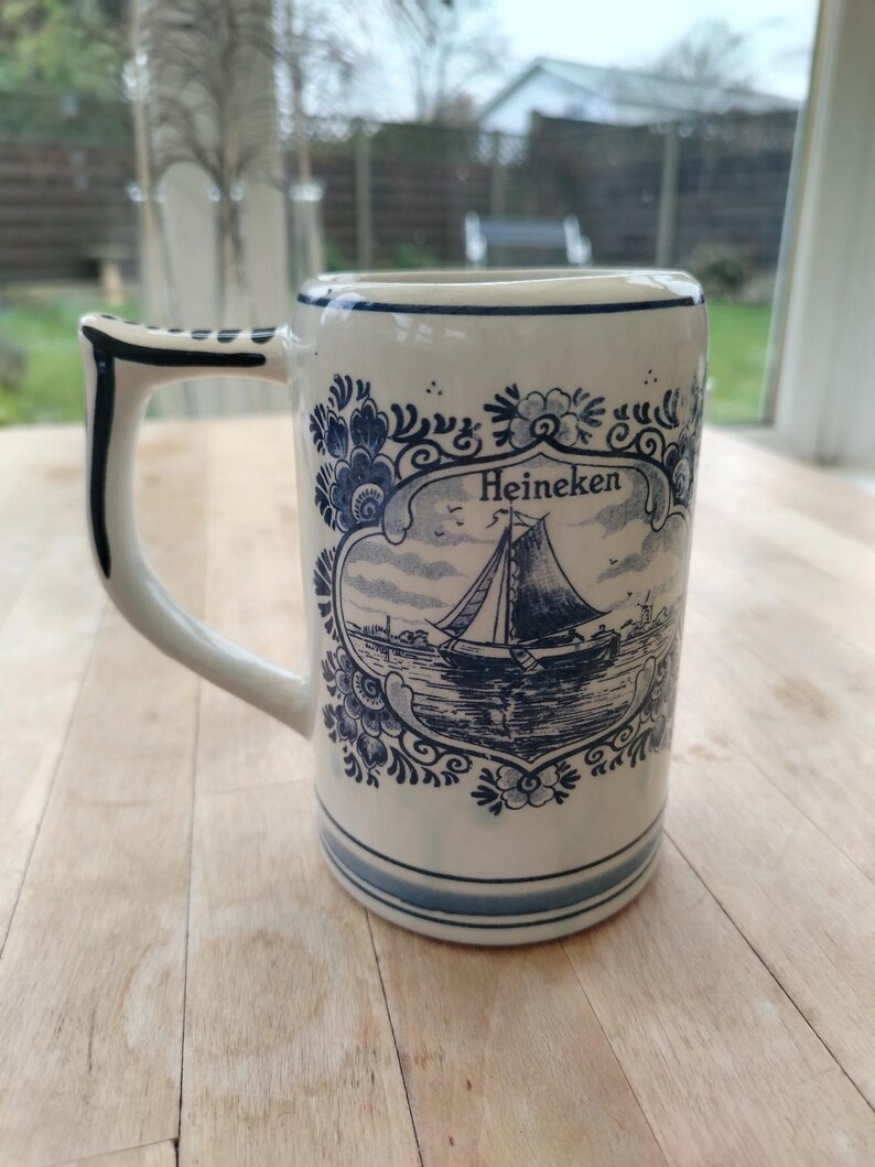 May include: A white ceramic beer stein with a blue and white floral design and the word "Heineken". The stein features a handle and a detailed illustration of a sailboat on the front. The stein is approximately 6 inches tall.