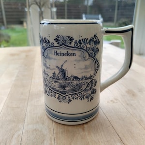May include: A white ceramic mug with a blue and white windmill design and the word "Heineken". The mug has a rectangular shape with a handle and blue accents. The design includes floral patterns.
