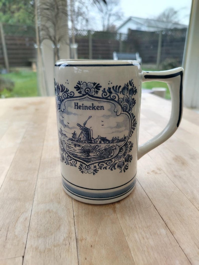 May include: A white ceramic mug with a blue and white windmill design and the word "Heineken". The mug has a rectangular shape with a handle and blue accents. The design includes floral patterns.