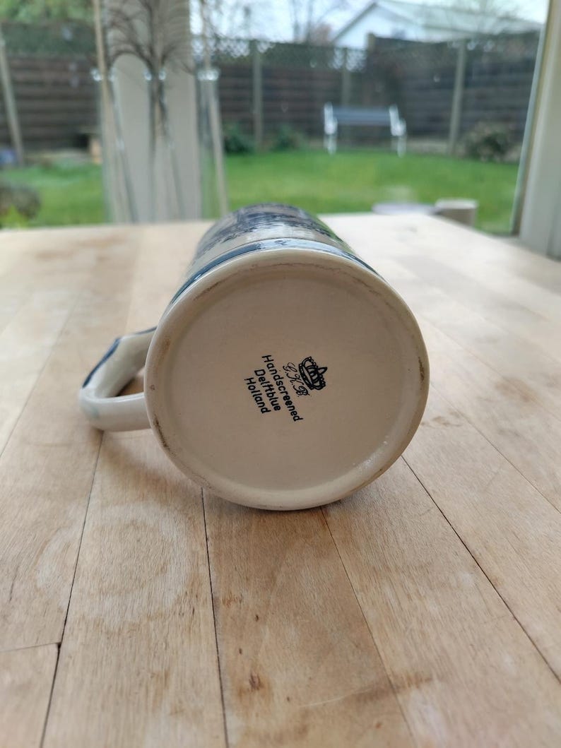 May include: A ceramic beer stein with a blue and white floral design. The stein has a handle and the text "Handscreened Delftblue Holland" is visible on the bottom. The stein is made of ceramic.