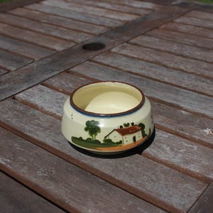 Puede incluir: Un cuenco pequeño y redondo de cerámica con interior crema y exterior pintado. El exterior presenta un diseño de paisaje con una casa, árboles y un borde azul. El cuenco está sobre una superficie de madera envejecida.