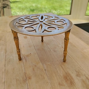 May include: A round brass trivet featuring an intricate openwork floral pattern on the top surface. The piece is supported by three slender, tapered legs with a decorative ringed design, providing a stable base for this metallic kitchen accessory.