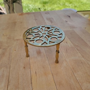 May include: A round brass trivet featuring an intricate openwork floral pattern on the top surface. The metal stand is supported by three slender, turned legs that elevate the circular plate, showcasing a polished metallic finish throughout.