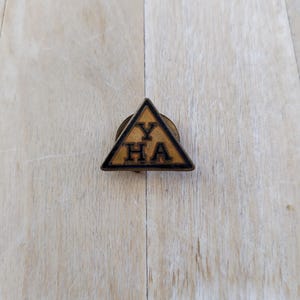 Könnte beinhalten: Ein goldfarbener dreieckiger Pin mit schwarzer Beschriftung, die "YHA" innerhalb eines Dreiecks lautet.