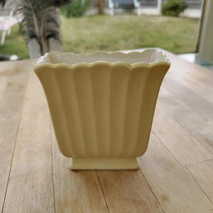 May include: A pale yellow ceramic vase with a square base and a flared, scalloped rim. The exterior features vertical fluted ridges running from the base to the top edge, creating a textured, pleated appearance with a smooth, glossy finish.