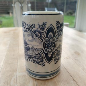 May include: A cylindrical ceramic mug with a cream-colored base and blue decorative patterns. The mug features a nautical scene with a sailboat and windmill, and the word "neken" is visible. The design is reminiscent of Delftware.
