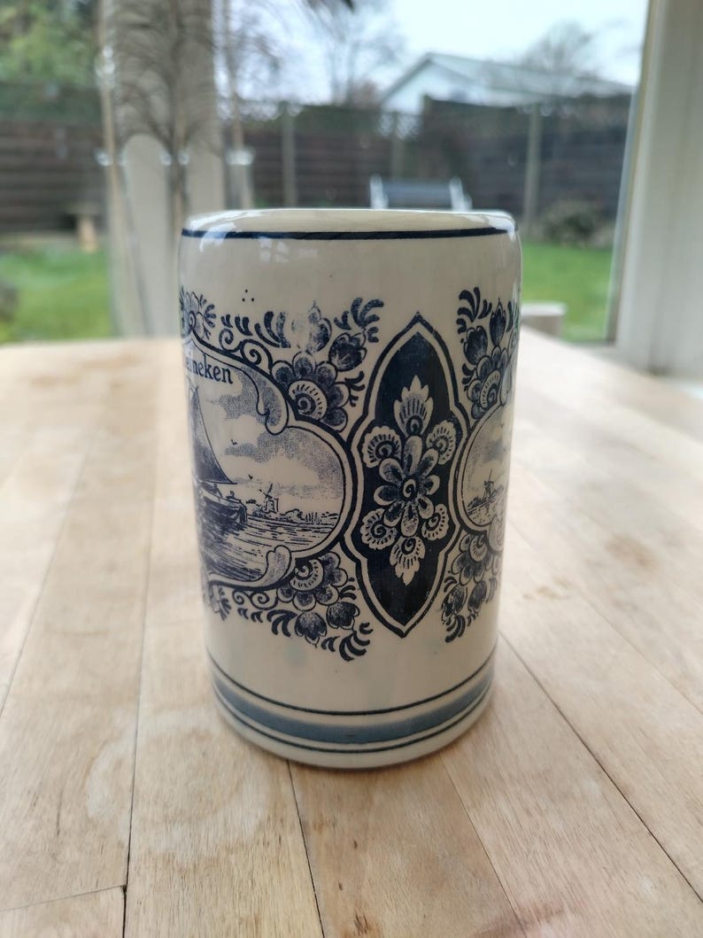 May include: A cylindrical ceramic mug with a cream-colored base and blue decorative patterns. The mug features a nautical scene with a sailboat and windmill, and the word "neken" is visible. The design is reminiscent of Delftware.