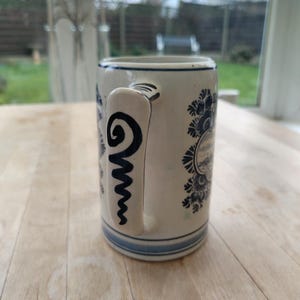 May include: A ceramic mug with a cream-colored base and blue floral and landscape designs. The handle features a black spiral design. The mug has a blue rim and base, and is likely used for drinking beverages.