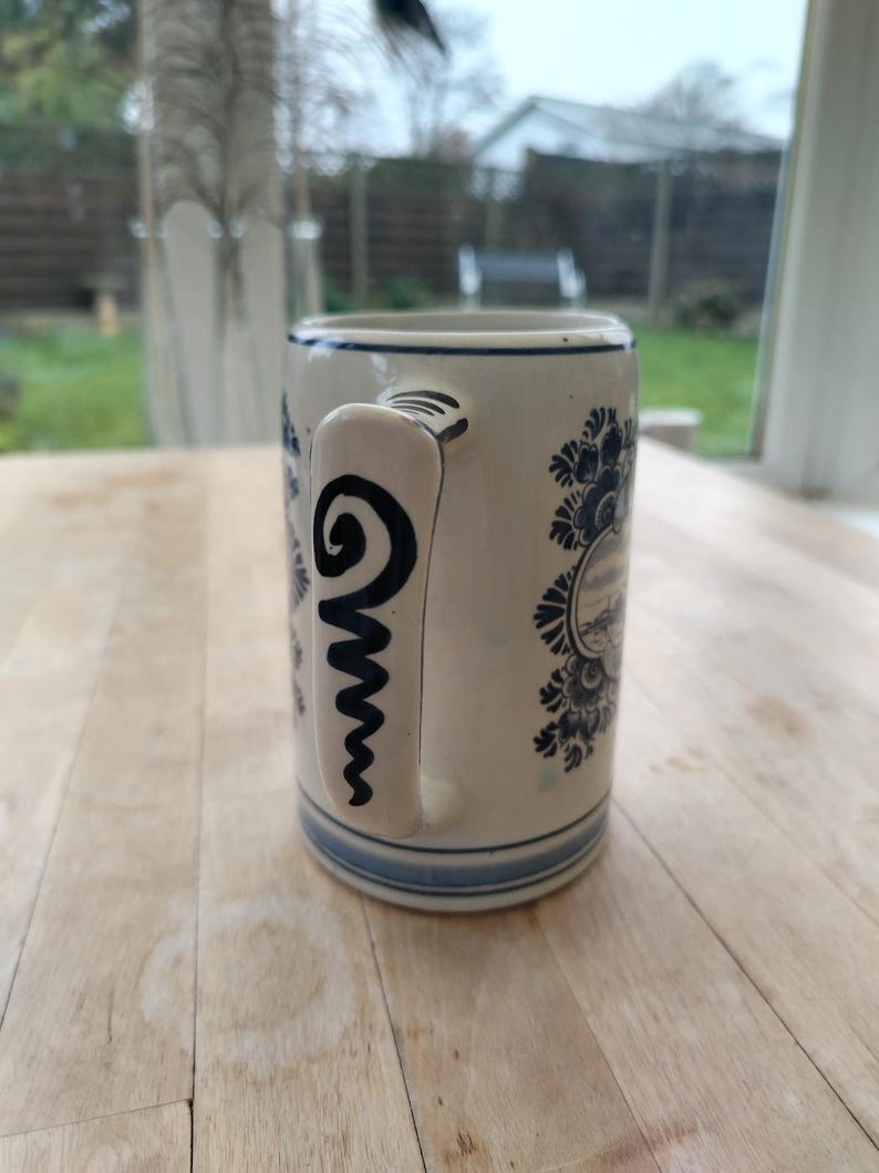 May include: A ceramic mug with a cream-colored base and blue floral and landscape designs. The handle features a black spiral design. The mug has a blue rim and base, and is likely used for drinking beverages.