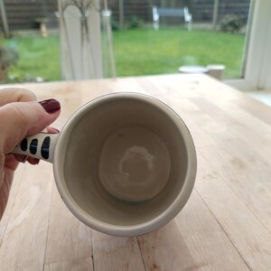 May include: A cream-colored ceramic mug with a black and white patterned handle. The mug's interior is a light beige color. The mug is held over a light-colored wooden surface, with a view of a backyard through a window in the background.