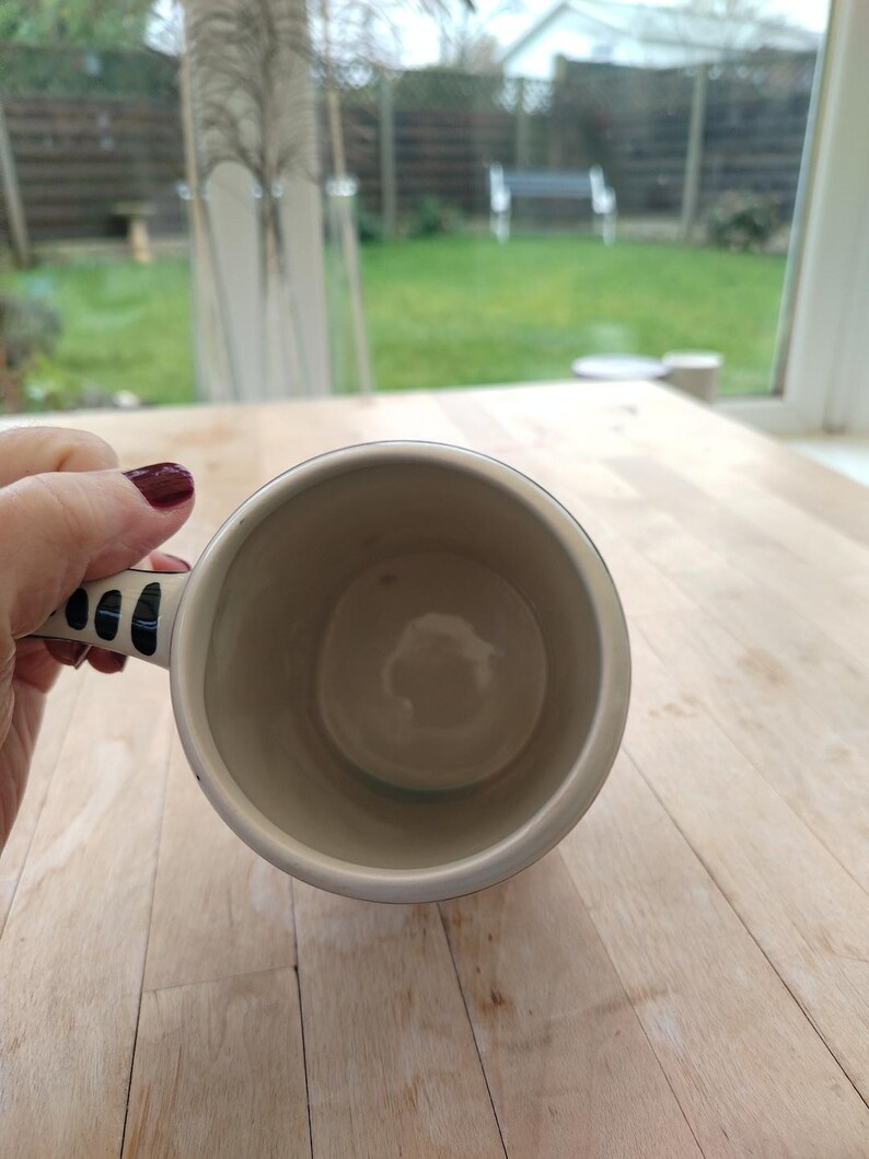 May include: A cream-colored ceramic mug with a black and white patterned handle. The mug's interior is a light beige color. The mug is held over a light-colored wooden surface, with a view of a backyard through a window in the background.