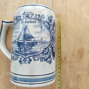 May include: A white ceramic beer stein with a blue and white sailboat design and the word "Heineken". The stein has a handle and blue stripes around the base. The stein is approximately 8 inches tall.