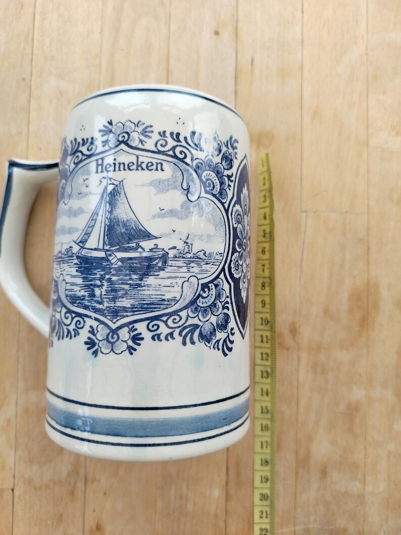 May include: A white ceramic beer stein with a blue and white sailboat design and the word "Heineken". The stein has a handle and blue stripes around the base. The stein is approximately 8 inches tall.